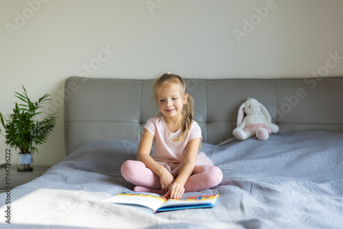 Young girl reading a book while sitting on a bed in a brightly lit bedroom. Peaceful morning moment