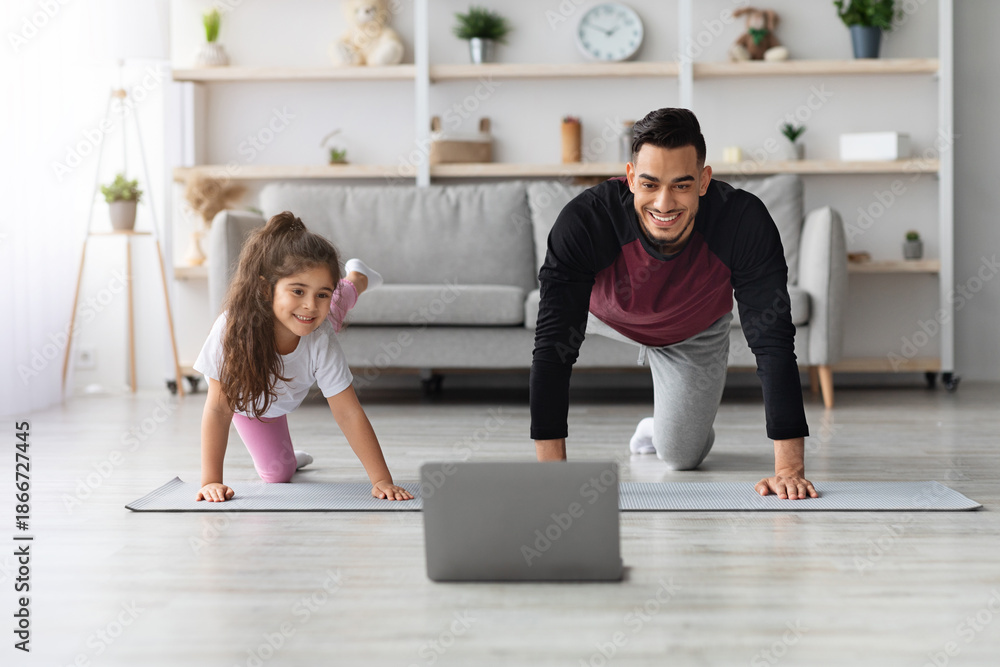 Obraz premium Happy arab father and little daughter having online fitness class, exercising at home together, wearing sportswear, training on yoga mat in front of laptop, enjoying workout, copy space