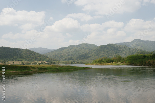 Beautiful outdoor view at Huai Tha Khoei Ban Kha Ratchaburi reservoir. The scene features a wooden photo spot, white ducks, and a calm lake with mountain views.