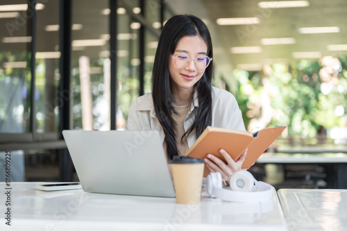 Glasses asian woman college student reading book aside laptop as sitting at white table in building Free Download Glasses asian woman college student reading book aside laptop as sitting at white table in building - GFXLoad
