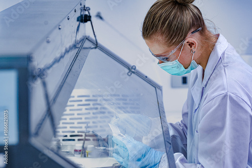 Slika na platnu Close-up of focused female scientist wearing mask and safety glasses working in sterile fume hood