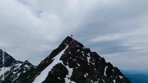 A craggy mountain peak with a red flag, snow patches under cloudy skies. Another mountain beside