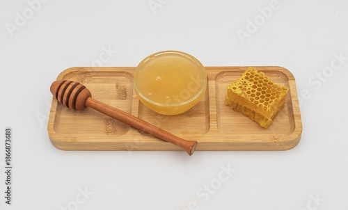 Glass bowl filled with honey, honeycomb piece and wooden dipper on bamboo tray. Clean white background, natural sweet food and organic ingredient concept.