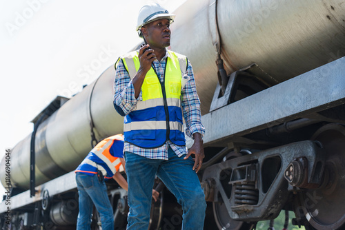 Industrial worker using walkie talkie during railway tanker inspection, Engineer inspecting freight train with safety helmet and vest, Railway maintenance worker communicating on site