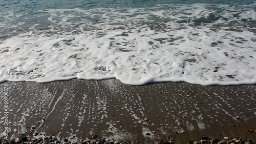 Gentle ocean wave with white foam washing over a dark pebble beach. Serene coastal landscape, sea water meeting the shore, natural textures of sand and stones.
