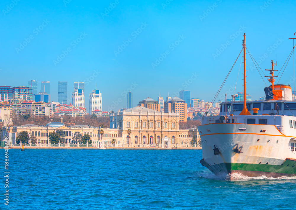 Fototapeta premium Sea voyage with old ferry (steamboat) in the Bosporus - Dolmabahce Palace seen from the Bosphorus - Dolmabahce palace against coastal cityscape with modern buildings under cloudy sky - Istanbul