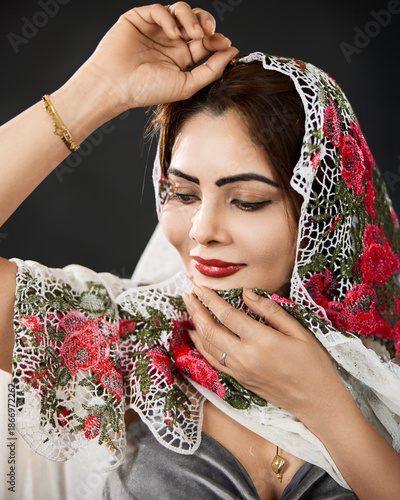 Close up studio portrait of a woman wearing a floral lace scarf and looking down against a dark background.