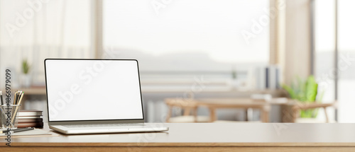 White screen laptop with books and pencils on wooden counter table across window wall in living room