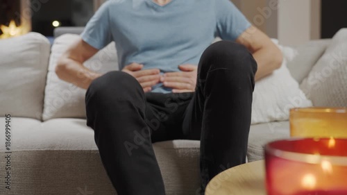 Man sitting on sofa in living room holding stomach and suffering from abdominal pain or cramps. Person suffering from health issues, digestion problems or food poisoning at home