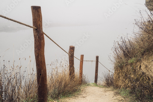 Foggy dirt trail with wooden posts and dry bushes on hillside during autumn morning