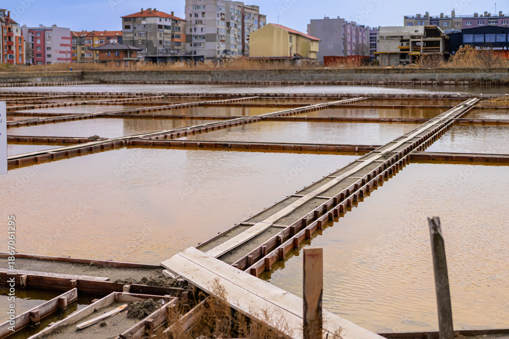 Fototapeta premium Salt evaporation ponds with wooden frames reflecting sky in Pomorie, Bulgaria, surrounded by residential buildings. Concept of traditional saltwork within modern coastal town