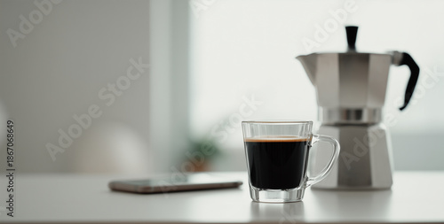 Glass cup of fresh black coffee with crema and a classic moka pot on a white table in a bright minimalist room, morning routine and workspace concept for home office productivity