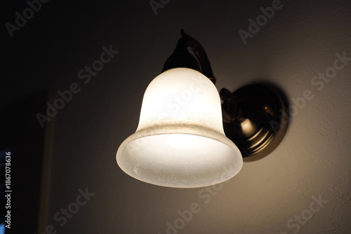 暗い部屋の壁に取り付けられたアンティーク調のウォールランプが温かい光を放っている室内風景です。磨りガラスのシェード越しに広がる柔らかな灯りが落ち着いた安らぎの空間を演出しています。