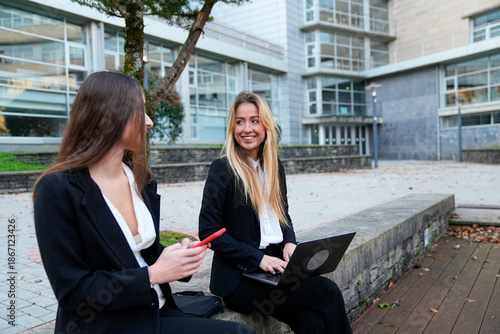Two young businesswomen collaborating and smiling, working outdoors with laptop and smartphone in an urban environment