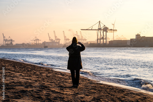 A person standing at the beach at the Elbe River in Hamburg taking photo of the sunrise over the harbor