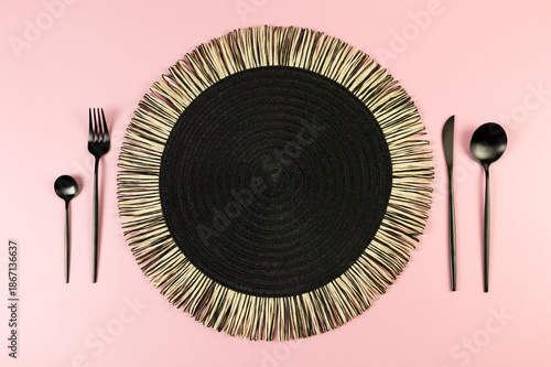 Top view of black wicker table mat, black cutlery on pink background. Straw round tray, flat lay, copy space. 