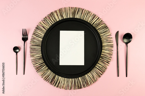 Top view of black wicker table mat, black cutlery, white card on pink background. Straw round tray, flat lay, copy space. 