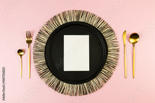 Top view of black wicker table mat, black plate, gold cutlery, white card on pink background. Straw round tray, flat lay, copy space. 