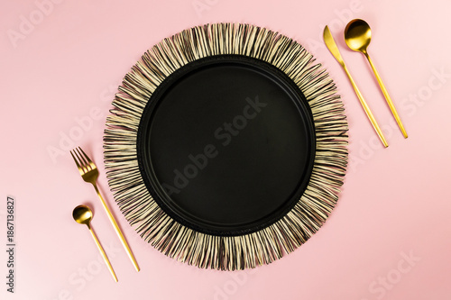 Top view of black wicker table mat, black plate, gold cutlery on pink background. Straw round tray, flat lay, copy space. 
