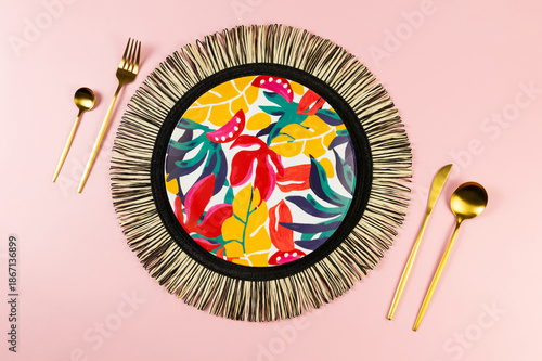 Top view of black wicker table mat, colorful plate, gold cutlery on pink background. Straw round tray, exotic plant plate, summer picnic. Flat lay, copy space. 