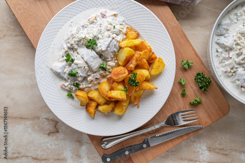 German homemade Herring salad with sour cream sauce, apples, pickles and onions. Served with fried potatoes on plate.