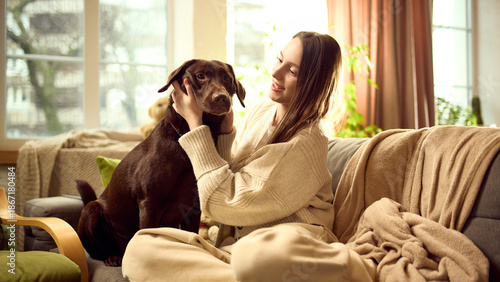 Dog sitting calmly on sofa at home while owner gently holds its head. Concept of pet care advertising, dog health check, everyday pet care routine and responsible home pet maintenance.