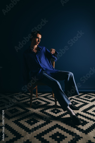 Foto Stylish man in elegant blue attire seated on chair against dark backdrop