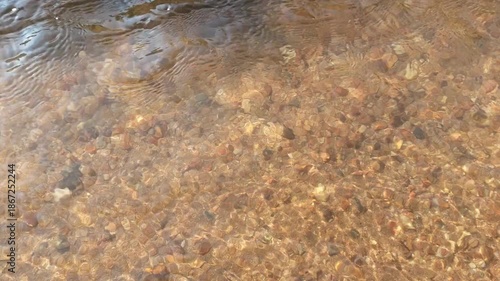 Peaceful clear shallow stream water flowing over brown stone pebble rock texture background showing natural tranquility of sand creek bed bottom with calm relaxing wave movement pure nature fresh