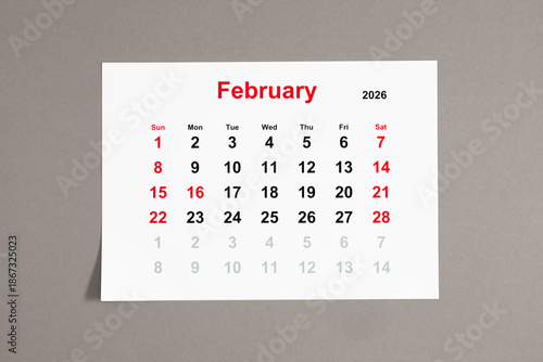 A close-up of a monthly tear-off calendar for February 2026 with weekends highlighted in red. The week starts on Sunday. Calendar sheet on a neutral gray background
