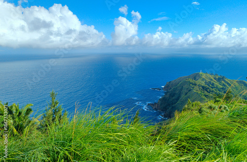 A scenic view captured from the top of a mountain, showcasing rolling green hills with grass and trees leading toward a vast blue ocean under a clear blue sky.