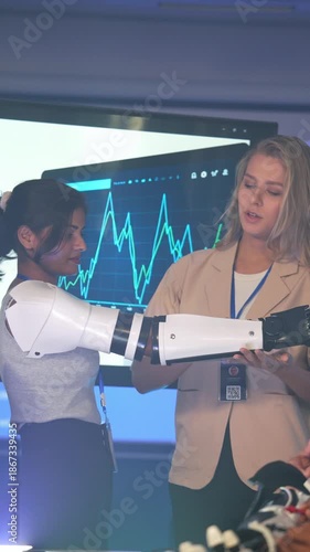 Female engineer explaining robotic prosthetic arm functions to a colleague wearing the device. Expert demonstration of assistive technology, AI rehabilitation, and bionic innovation for disability.