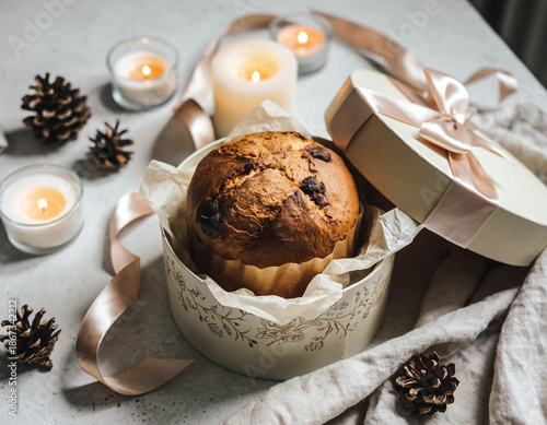 Festive panettone cake in elegant gift box with ribbon, surrounded by candles and pinecones for cozy holiday or Christmas celebration