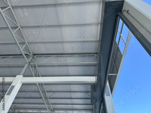 Tableau sur toile Interior view of a modern warehouse roof featuring steel trusses, insulated metal panels, and structural beams, captured from a low angle with blue sky visible
