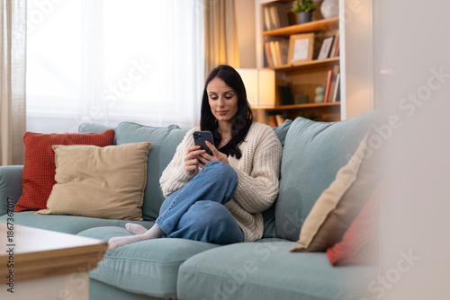 Young woman using a smartphone while sitting on a sofa at home. Calm lifestyle scene with cozy living room, natural light, and copy space.
