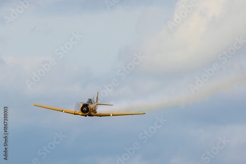 Canadian Car and Foundry Harvard Mk.IV aircraft flying with smoke trail against cloudy sky.