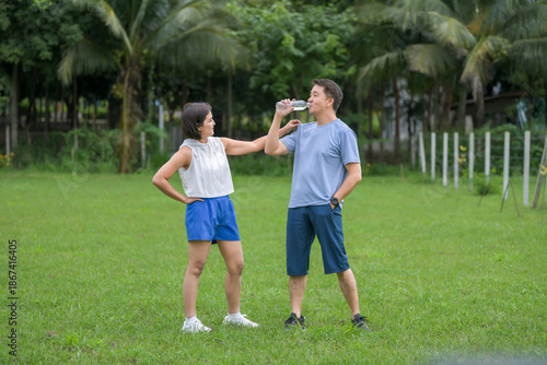 A man and woman in athletic wear standing on a green field; the man is drinking water from a bottle while the woman stands with her hands on her hips.