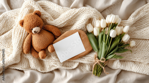 Teddy bear and white tulips with blank card on a cozy blanket. A charming scene of a teddy bear, flowers, and a blank card creates a warm and inviting atmosphere