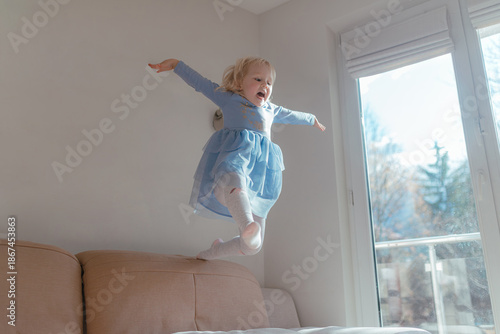 Fotografi Young girl descends toward sofa wearing layered blue dress, arms open wide, brig