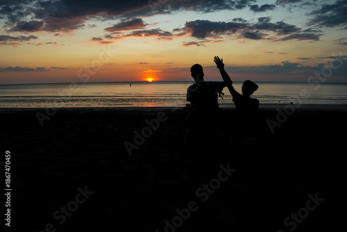 Silhouette of a wheelchair user and caregiver watching the sunset at the beach, hands raised with happiness, symbol of freedom, inclusion, accessibility and positive life attitude.