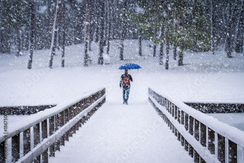 Man walking alone across a bridge with umbrella during winter snowfall. Cold weather, daily commute, loneliness and winter lifestyle concept.