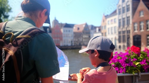 Family tourists using a map together, standing on a bridge by a canal with charming old buildings and colorful flowers, enjoying a lively summer city vacation