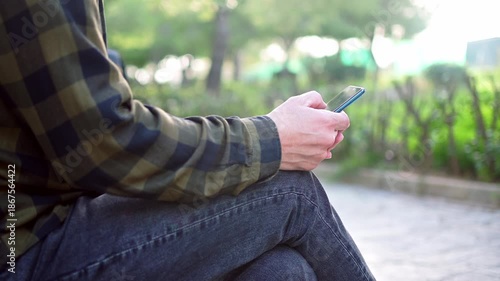 Vertical footage shows a man wearing gray jeans and plaid green shirt using his mobile phone outdoors. Selective focus, nice soft background.