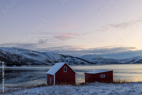 Zwei kleine norwegische Holzhütten am ruhigen See in winterlicher Landschaft, schneebedeckte Ufer und Berge im Hintergrund, klare Abendstimmung mit sanftem Restlicht am Himmel 