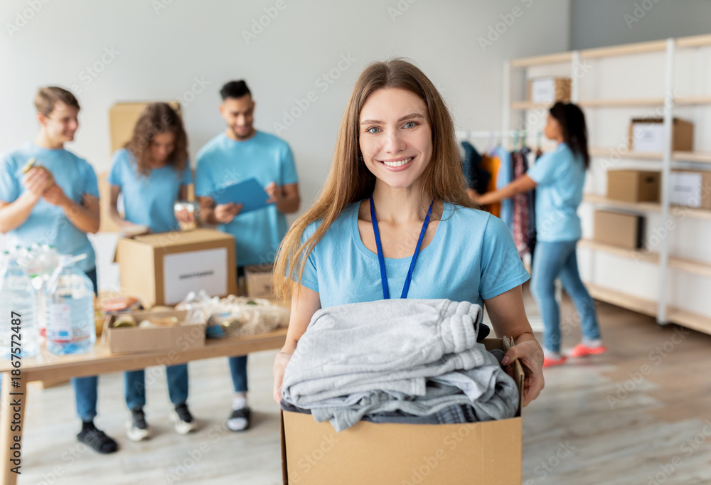 Obraz premium A cheerful female volunteer smiles while holding a box of clothes for donation. She works alongside other young volunteers, packing items together at the charity center, fostering community spirit.