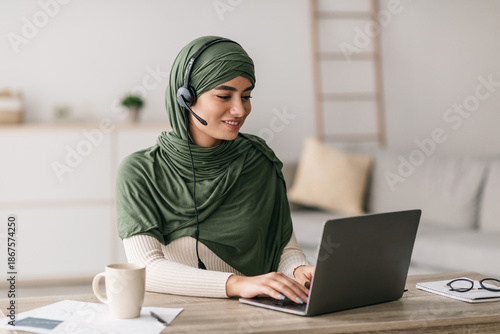 A millennial Arab lady in a green hijab is using her laptop and wearing headphones. She is working remotely from home and actively communicating with a client during an online meeting.