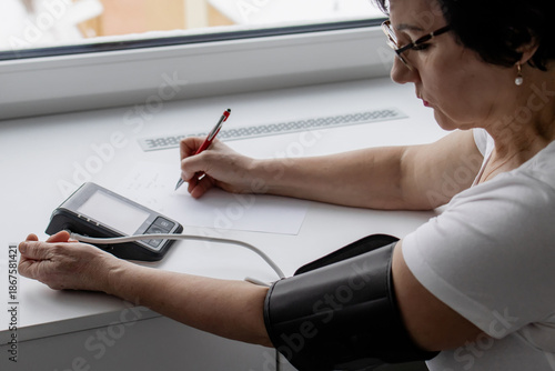 Senior woman measuring blood pressure at home and writing results on paper while sitting by the window during winter, self-monitoring health and managing hypertension