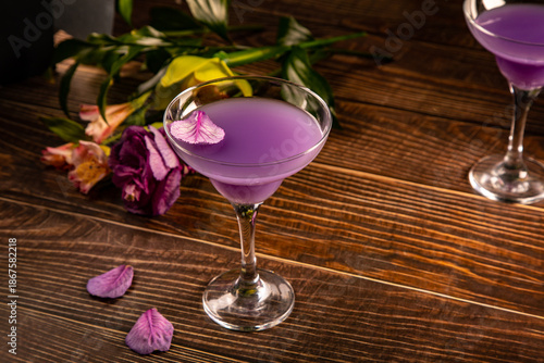 Elegant martini glass filled with a vibrant purple cocktail, placed on a wooden surface with scattered purple petals and a blurred floral background.