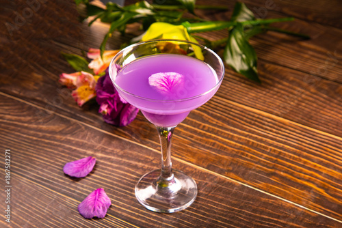 Elegant martini glass filled with a vibrant purple cocktail, placed on a wooden surface with scattered purple petals and a blurred floral background.