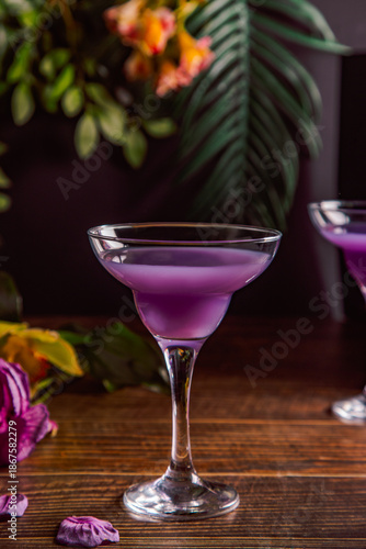 Elegant martini glass filled with a vibrant purple cocktail, placed on a wooden surface with scattered purple petals and a blurred floral tropical background.