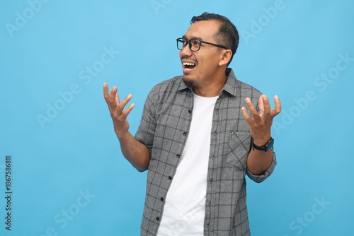 Fényképezés Asian man with confused expression raising his hands, shrugging his shoulders in confusion, not knowing what to do, wearing casual shirt, isolated on blue studio background